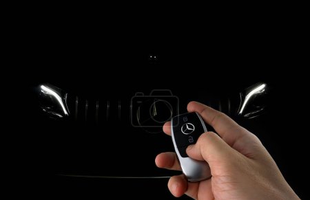 Photo for Right hand holding car remote key to unlock car Benz c200 coupe AMG dynamic model with background of Mercedes Benz chromium grille star logo and led headlight park in the dark garage - Royalty Free Image