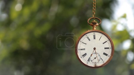 Photo for Pocket Watch on Chain Time is Ticking features a gold pocket watch hanging from a gold chain with blurred leaves blowing in the wind in the background. - Royalty Free Image