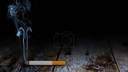 Photo for Smoke of Death Table Top Background features a cigarette laying on a rustic wooden table top with smoke rising from the tip of the cigarette. - Royalty Free Image