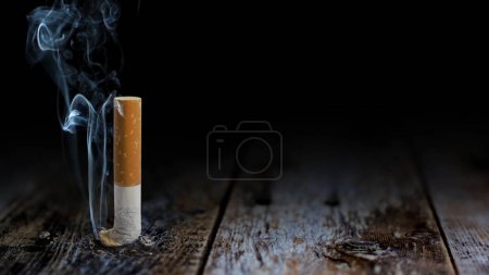 Photo for Smoke of Death Table Butt Background features a cigarette crushed out on a rustic wooden table top with smoke rising from the tip of the cigarette. - Royalty Free Image