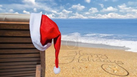 Photo for Happy Holidays from the Beach Santa Hat features a beach chair with a Santa hat hanging with an ocean in the background and Happy Holidays written in the sand, Not A.I. generated. - Royalty Free Image