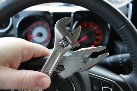 Zangen und einstellbarer Schraubenschlüssel in menschlichen Fingern vor defokussiertem Auto-Armaturenbrett mit roten Ziffern und Pfeilen  