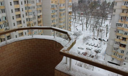 View from a snow-covered balcony of cars under snow parked near multi-storey buildings