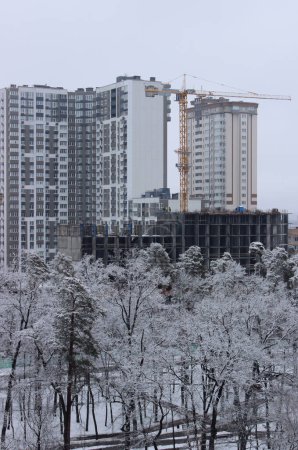Grue à haute altitude sur le chantier de construction d'un bâtiment moderne multi-étages derrière un petit parc municipal couvert de neige 