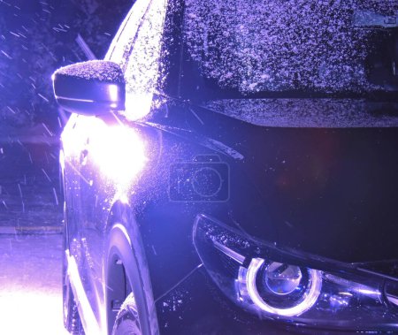 Winternacht fahren Archivfoto. Reflexion von Autoscheinwerfern an der Tür eines am Straßenrand geparkten schwarzen Autos bei nächtlichem Schneefall