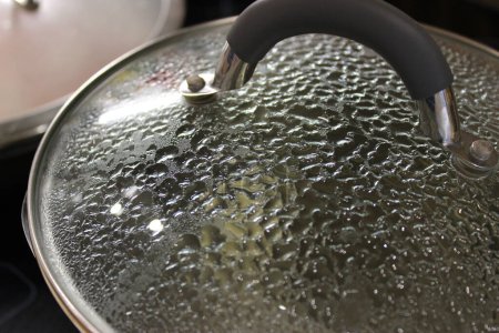Steam turns into water droplets on the inside of a glass lid of a boiling pan