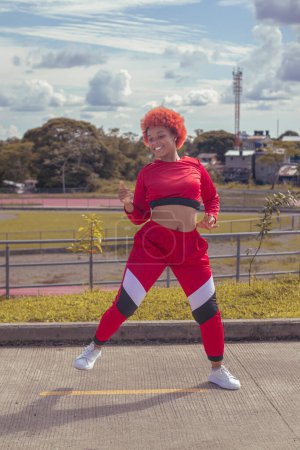 Photo for Joven afro de cabello naranja bailando al estilo urbano en parque local con pista de atletismo, vegetacion y cielo hermosos de fondo - Royalty Free Image