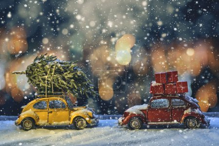 Photo for Two models of old passenger cars with a Christmas tree and gifts on the roof stand under the snow in the evening electric light. - Royalty Free Image