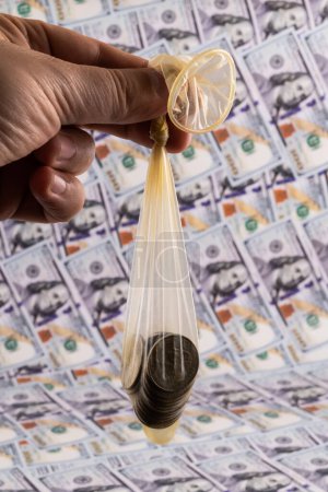 Photo for A condom filled with 25 American cents in a man's hand against the background of 100 American dollars bills - Royalty Free Image