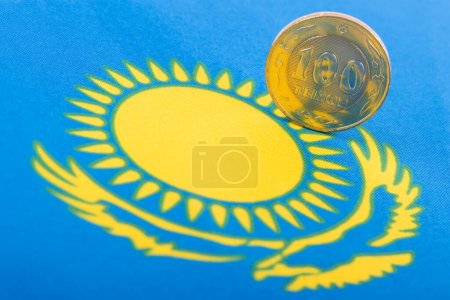 Moneda en denominación de 100 tenge kazajo sobre el fondo de un fragmento de la bandera kazaja con un águila voladora y el Sol