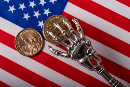 Foto de Mano humana esqueletizada de metal y monedas en denominaciones de 1 dólar americano en el fondo de la bandera americana - Imagen libre de derechos
