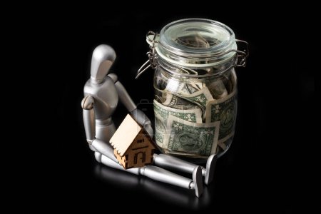 Téléchargez les photos : Pot en verre avec billets de 1 dollar, homme en bois et maison symbolique sur fond noir - en image libre de droit