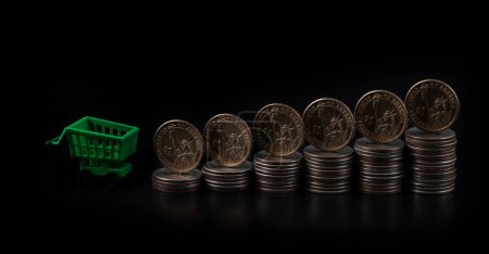 Foto de Monedas de 1 dólar y carrito de compras de supermercado en miniatura sobre fondo negro - Imagen libre de derechos