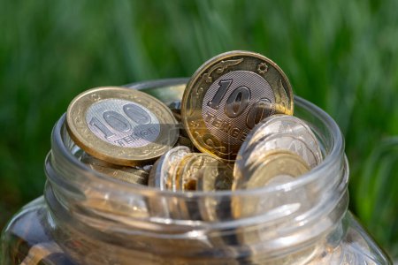 Coins of 100 and 200 Kazakhstani tenge in a glass jar on a background of grass