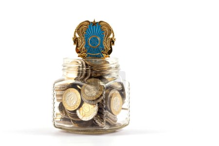 Coat of arms of the Republic of Kazakhstan and coins of 100 and 200 Kazakhstan tenge in a glass jar on a white background