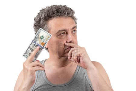 Foto de Un hombre de mediana edad peludo y canoso con rastrojo y una camiseta sin mangas tiene un billete de 100 dólares en sus manos. - Imagen libre de derechos