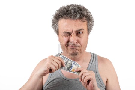 Un hombre de mediana edad peludo y canoso con rastrojo y una camiseta sin mangas tiene un billete de 100 dólares en sus manos.