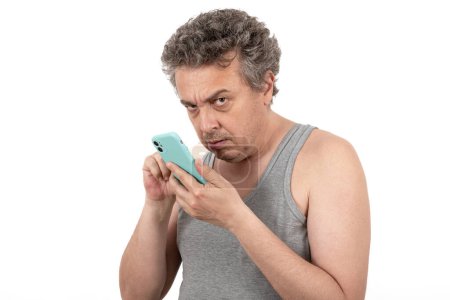 A gray-haired, shaggy, unshaven middle-aged man in a sleeveless T-shirt holds a smartphone