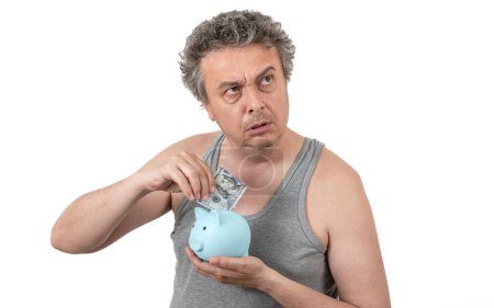 Ein grauhaariger, zotteliger, unrasierter Mann mittleren Alters in einem ärmellosen T-Shirt hält ein Sparschwein und einen 100-Dollar-Schein in den Händen..