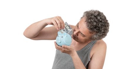 A gray-haired, shaggy, unshaven middle-aged man in a sleeveless T-shirt holds a piggy bank and a 100 US dollar bill in his hands.