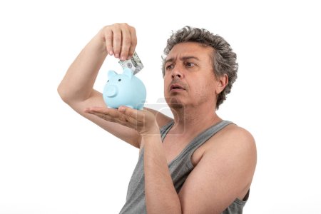 Ein grauhaariger, zotteliger, unrasierter Mann mittleren Alters in einem ärmellosen T-Shirt hält ein Sparschwein und einen 100-Dollar-Schein in den Händen..