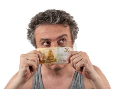 Un homme d'âge moyen aux cheveux gris, shaggy et non rasé dans un T-shirt sans manches tient un billet de 100 roubles russes