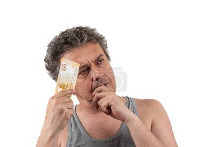 Un homme d'âge moyen aux cheveux gris, shaggy et non rasé dans un T-shirt sans manches tient un billet de 100 roubles russes