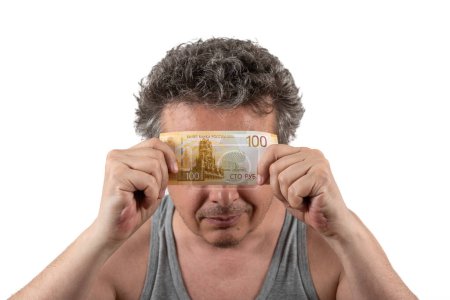 A gray-haired, shaggy, unshaven middle-aged man in a sleeveless T-shirt holds a 100 Russian ruble bill