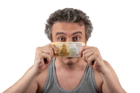 Un hombre de mediana edad, de pelo gris, peludo y sin afeitar, con una camiseta sin mangas, sostiene un billete de 100 rublos rusos.