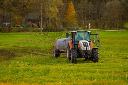 Photo for Modern tractor spreading manure on a green field before the rain.  Forest in the background. - Royalty Free Image