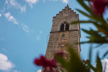 Foto de Campanario del centro de la ciudad de vipitieno, escondido detrás de algunas flores y frente a un hermoso cielo azul. Torre hecha de piedras cuadradas. - Imagen libre de derechos