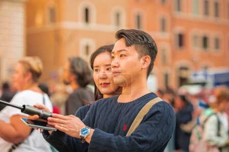 Foto de ROMA, ITALIA, 31.10.2023: Una pareja asiática se hace una selfie usando un bastón de selfie frente al monumento torcido de la capilla Sixtina en el centro de Roma. - Imagen libre de derechos