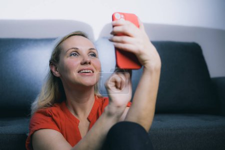 Young lady, woman in a red dress with a matching smart phone is taking a selfie. Mature online dating, long distance call, video call, photographing yourself. Frontal camera.