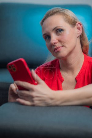 Young lady, woman in a red dress with a matching smart phone is taking a selfie. Mature online dating, long distance call, video call, photographing yourself. Looking at the camera.