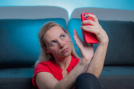 Junge Frau in rotem Kleid mit passendem Smartphone macht ein Selfie. Reifes Online-Dating, Ferngespräche, Videoanrufe, sich selbst fotografieren. Frontkamera.