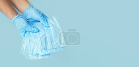 Foto de Hand in latex medical gloves and surgical ear-loop mask on blue background. Protection concept. copy space - Imagen libre de derechos