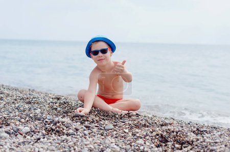 Foto de Pequeño niño feliz con gafas de sol azules y sombrero tiene una gran sonrisa en su cara. Cielo azul de verano y fondo de agua de mar. Niño en vacaciones de verano. - Imagen libre de derechos