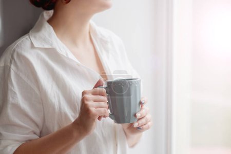 Photo for Closeup of female hands with a mug of beverage. Beautiful girl in grey sweater holding cup of tea or coffee in the morning sunlight. - Royalty Free Image