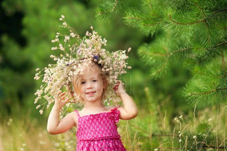 Téléchargez les photos : Little girl is wearing flower wreath on her head in field on summer sunny day. Portrait of adorable little child outdoors. happy holiday childhood. - en image libre de droit