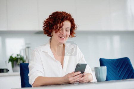 Téléchargez les photos : Femme heureuse utilisant un téléphone portable tout en travaillant à la maison. Messagerie femme souriante avec smartphone. Belle dame site de navigation sur téléphone portable - en image libre de droit