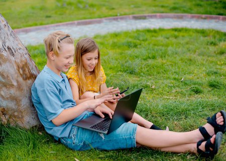 Téléchargez les photos : Deux adolescents heureux à la recherche de contenu multimédia en ligne dans un ordinateur portable assis sur l'herbe dans un parc - en image libre de droit