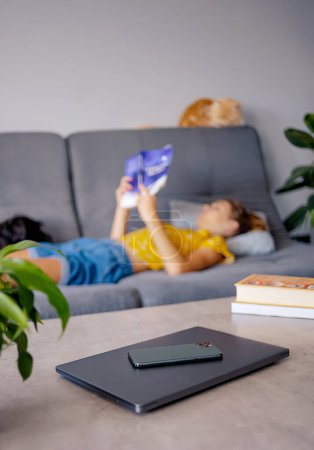 Téléchargez les photos : Concept numérique de désintoxication et de loisirs - gros plan des gadgets dans le panier sur la table et la fille lecture livre à la maison - en image libre de droit