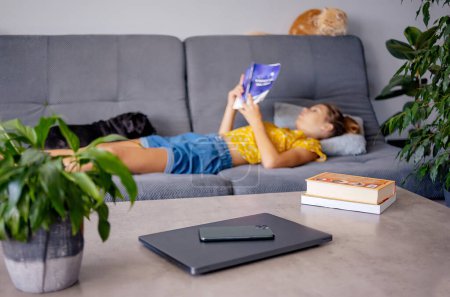 Téléchargez les photos : Concept numérique de désintoxication et de loisirs - gros plan des gadgets dans le panier sur la table et la fille lecture livre à la maison - en image libre de droit