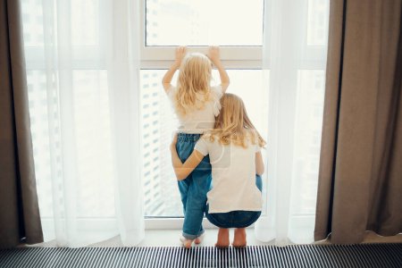 Téléchargez les photos : Les petites filles regardent par la fenêtre. les enfants regardent par la fenêtre. quarantaine. enseignement à distance - en image libre de droit