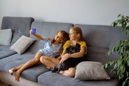 Téléchargez les photos : Filles assises sur le canapé, s'amuser à la maison, utiliser un smartphone et rire, jouer au jeu vidéo sur téléphone mobile, copier l'espace. concept enfants et gadgets - en image libre de droit