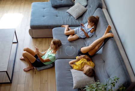 Foto de Niñas jugando juego móvil en el teléfono inteligente sentado en un sofá. Ocio infantil en casa, adicción a los videojuegos - Imagen libre de derechos