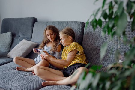 Téléchargez les photos : Filles jouant jeu mobile sur smartphone assis sur un canapé. Loisirs pour enfants à la maison, dépendance au jeu vidéo - en image libre de droit