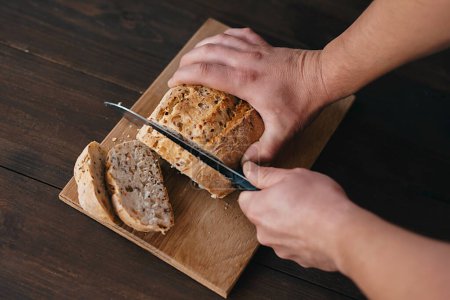 Photo for A person is slicing a bread loaf on a wooden cutting board. The staple food ingredient is a key component in various cuisine recipes - Royalty Free Image