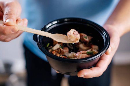 person is delicately holding a bowl of comfort food with a fork in it, possibly a warm dish made with seafood ingredients. take away