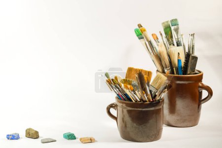 Photo for Artistic brushes, colorful nice stones and painting tools in old ceramic pots on a white background. Artistic composition. - Royalty Free Image